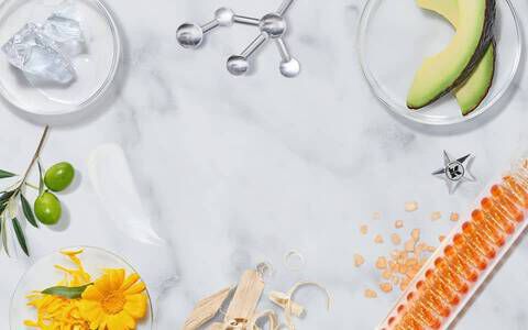 Disposizione di ingredienti naturali e attrezzature scientifiche su un piano di marmo, inclusi fiori, foglie, oli e piccole molecole.